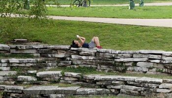 student lying on her back on stone wall reading a book against a green field with bicycles in the background